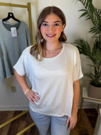 A woman modeling a short-sleeve ribbed knit top in cream/off-white, paired with light blue jeans, standing in a boutique setting with a clothing rack and potted plant in the background.