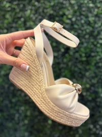 Beige wedge sandal with espadrille sole held against a green leafy background