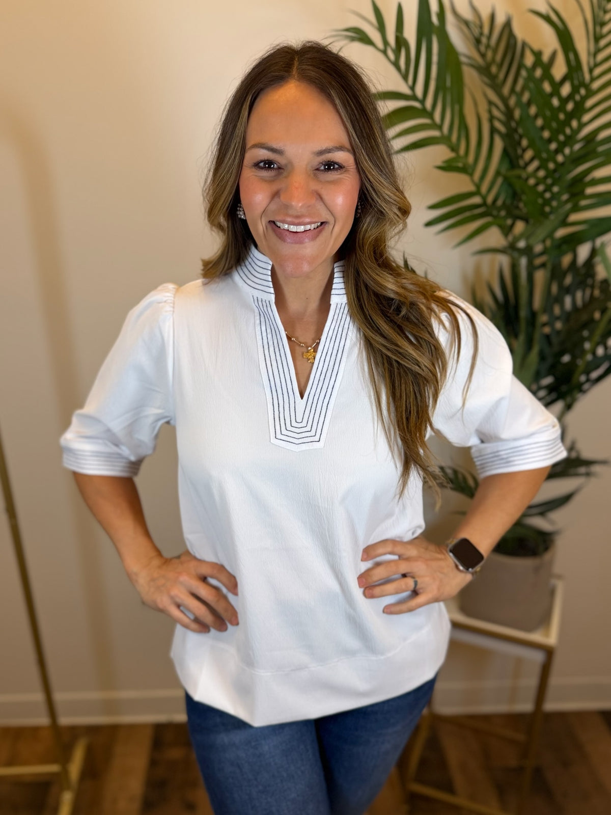 Woman wearing a white blouse with contrast stitching standing in a room with a plant in the background