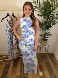 Woman wearing a white and blue floral maxi dress with ruching standing in a room with a rack of dresses and a plant.