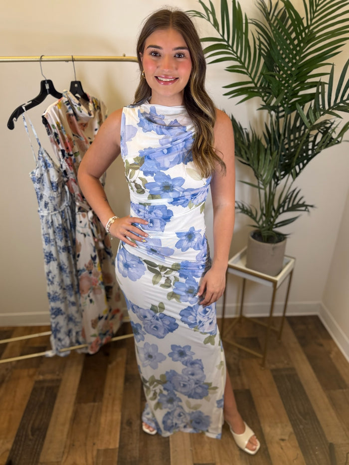Woman wearing a white and blue floral maxi dress with ruching standing in a room with a rack of dresses and a plant.