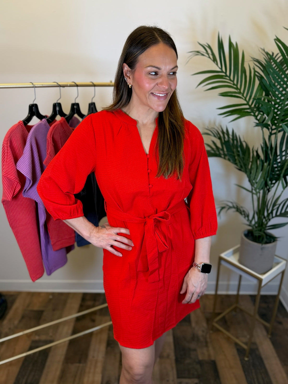 A woman modeling a bright red short dress with three-quarter sleeves and a tie waist, standing in a boutique setting with a clothing rack and potted plant in the background.