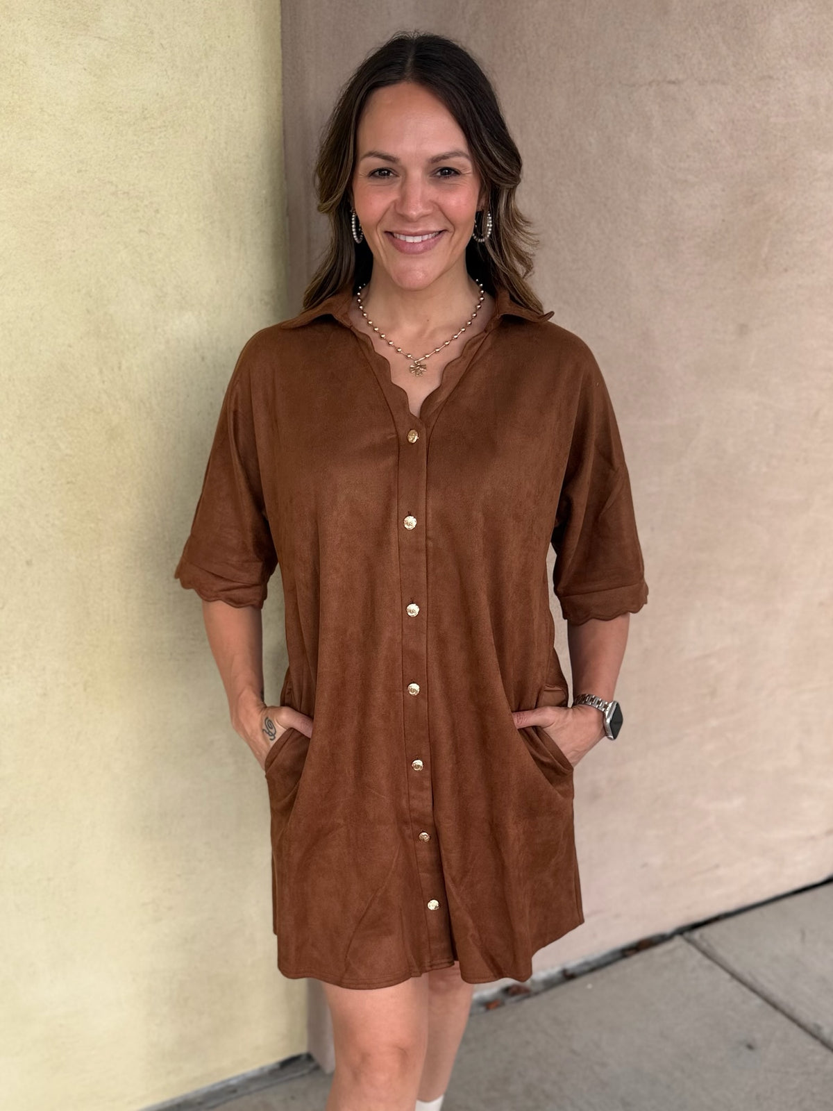 A woman wearing a short-sleeve camel button-front shirt dress with scalloped trim and pockets, standing outdoors against a beige stucco wall.