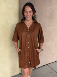 A woman wearing a short-sleeve camel button-front shirt dress with scalloped trim and pockets, standing outdoors against a beige stucco wall.