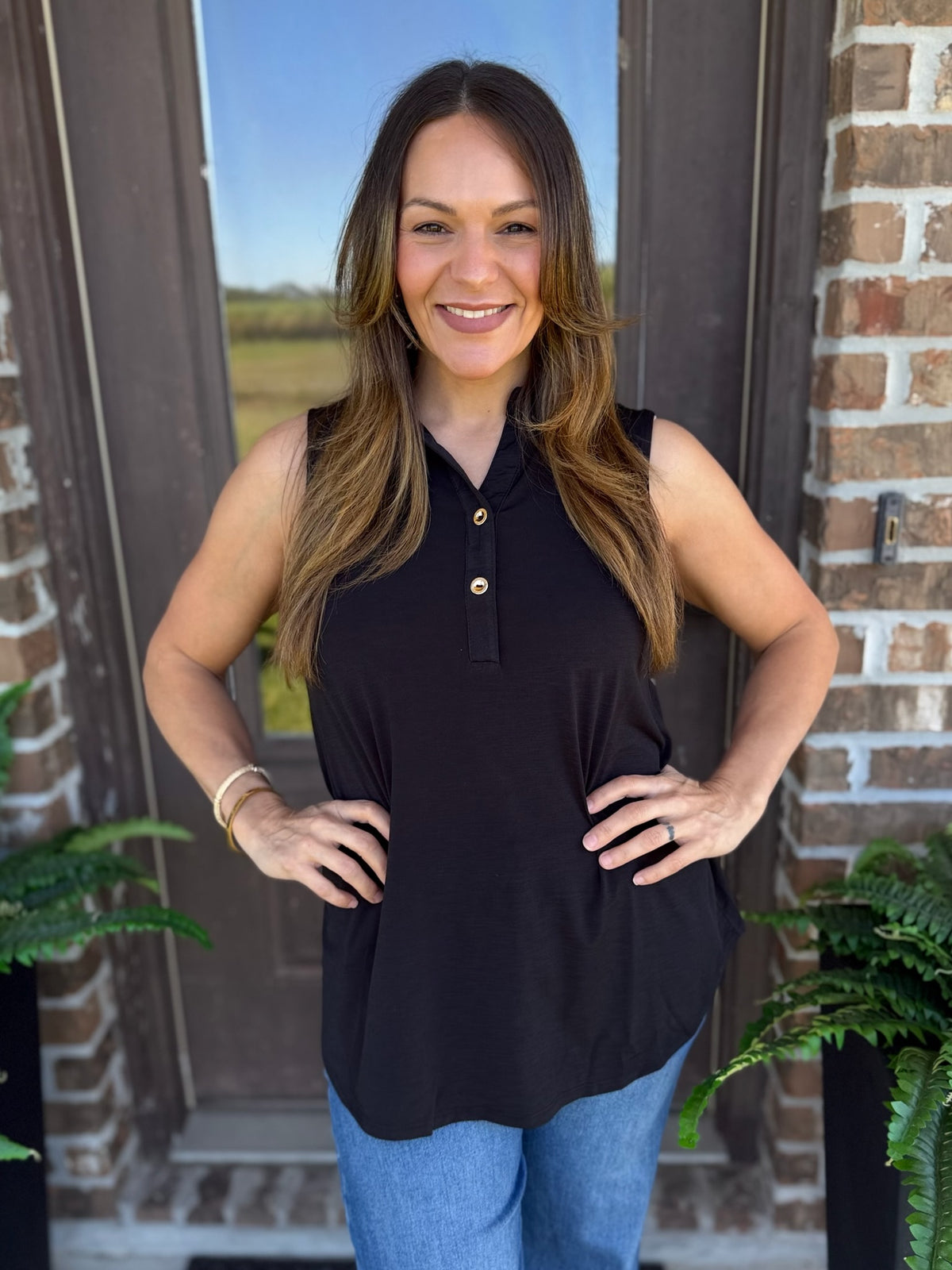 A woman with long brown hair stands outdoors in front of a brick door wearing a sleeveless black collared top with a small button placket and blue jeans.