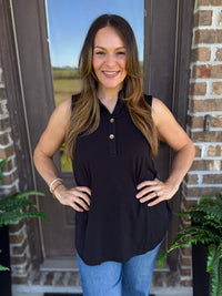 A woman with long brown hair stands outdoors in front of a brick door wearing a sleeveless black collared top with a small button placket and blue jeans.