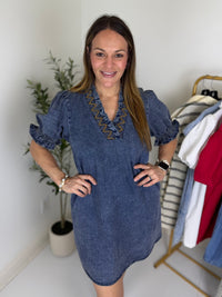 Woman wearing a dark blue denim dress with embroidered neckline and short puff sleeves in a room with a plant and clothing rack.