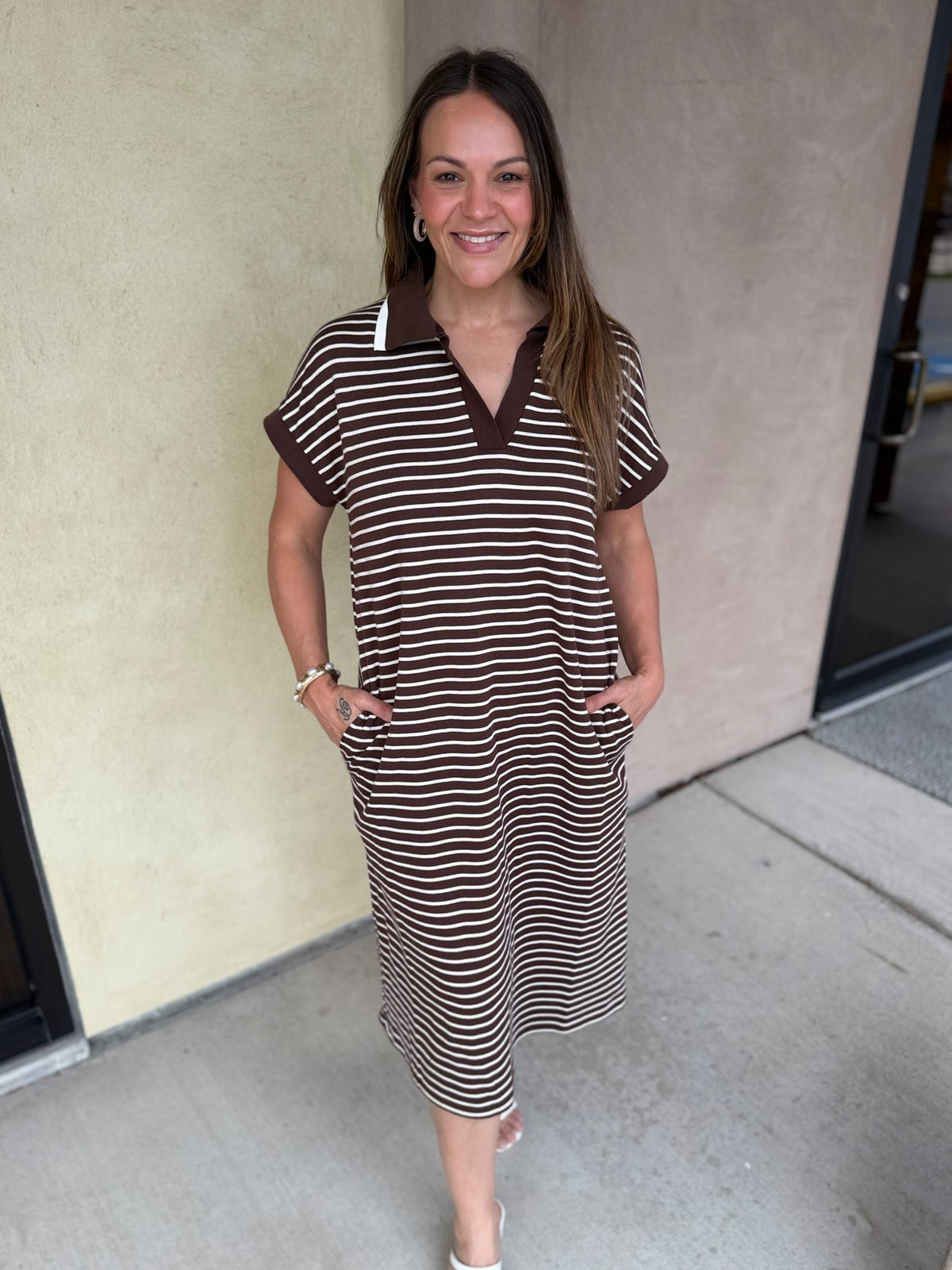 A woman modeling a brown and beige horizontally striped short-sleeve polo dress with a solid brown collar, standing outdoors against a beige stucco wall.