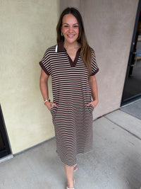 A woman modeling a brown and beige horizontally striped short-sleeve polo dress with a solid brown collar, standing outdoors against a beige stucco wall.