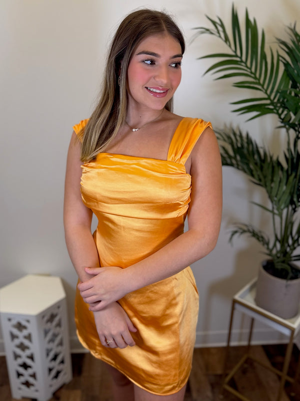 A woman wearing a shiny orange satin mini dress with a square neckline and ruched bodice, standing in an indoor setting with a potted plant and white furniture in the background.