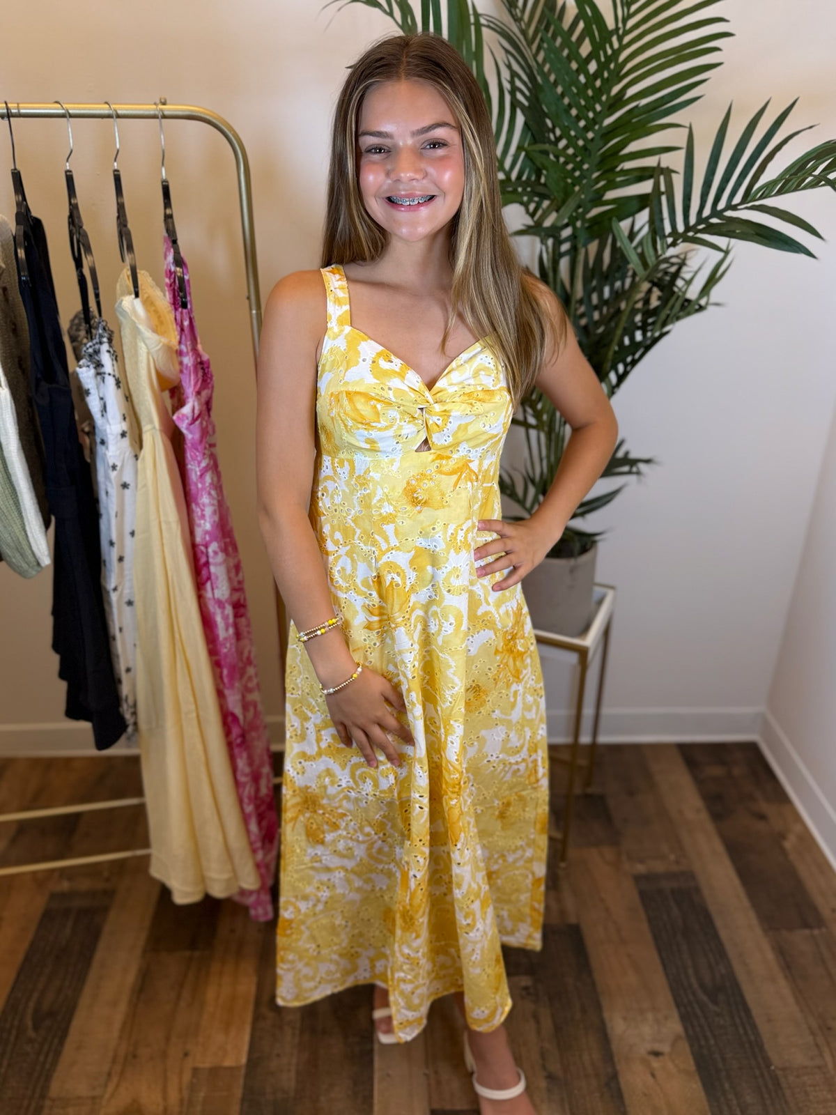 Woman wearing a sleeveless yellow floral dress in a room with a rack of dresses and a plant.