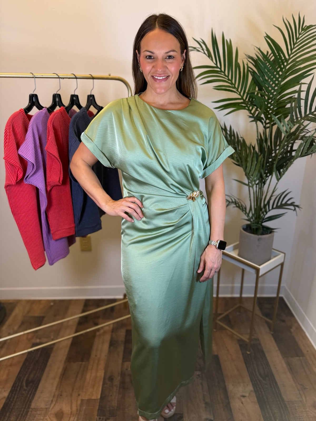 A woman modeling a sage green satin midi dress with short sleeves and a draped waist detail, standing in a boutique setting with a clothing rack and a potted plant in the background.