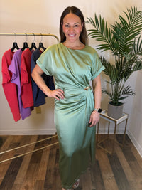 A woman modeling a sage green satin midi dress with short sleeves and a draped waist detail, standing in a boutique setting with a clothing rack and a potted plant in the background.