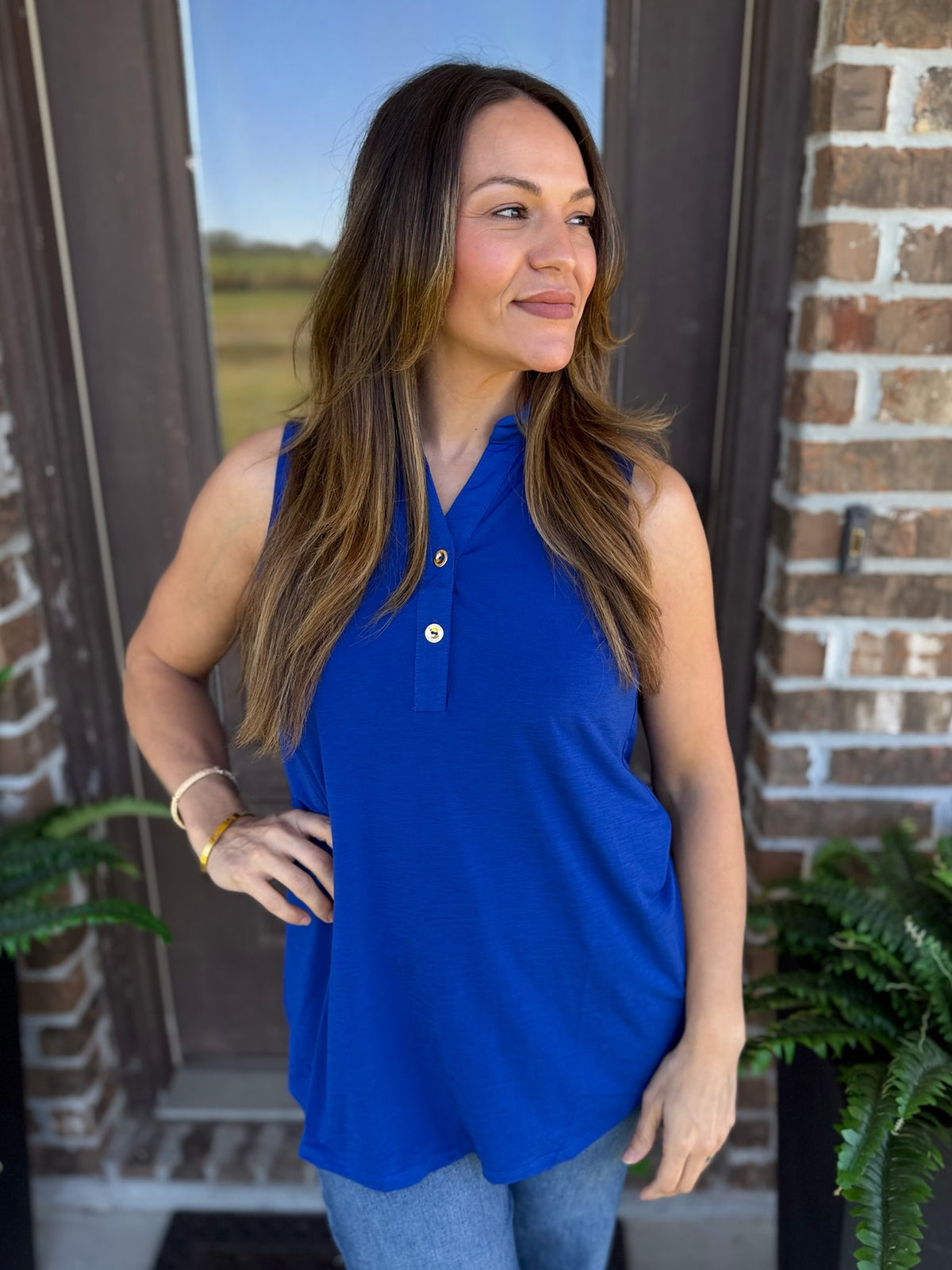 A woman wearing a sleeveless royal blue polo-style top with a small button placket, standing outdoors in front of a brick door with plants visible on both sides.