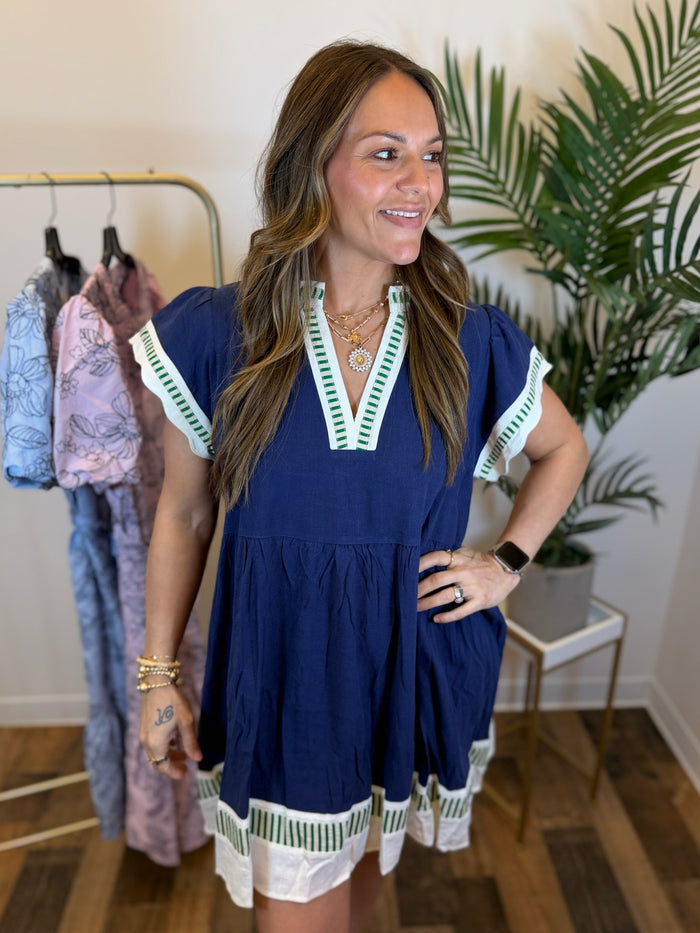 Woman wearing a short navy dress with white and green trim details and split neckline in a clothing store.