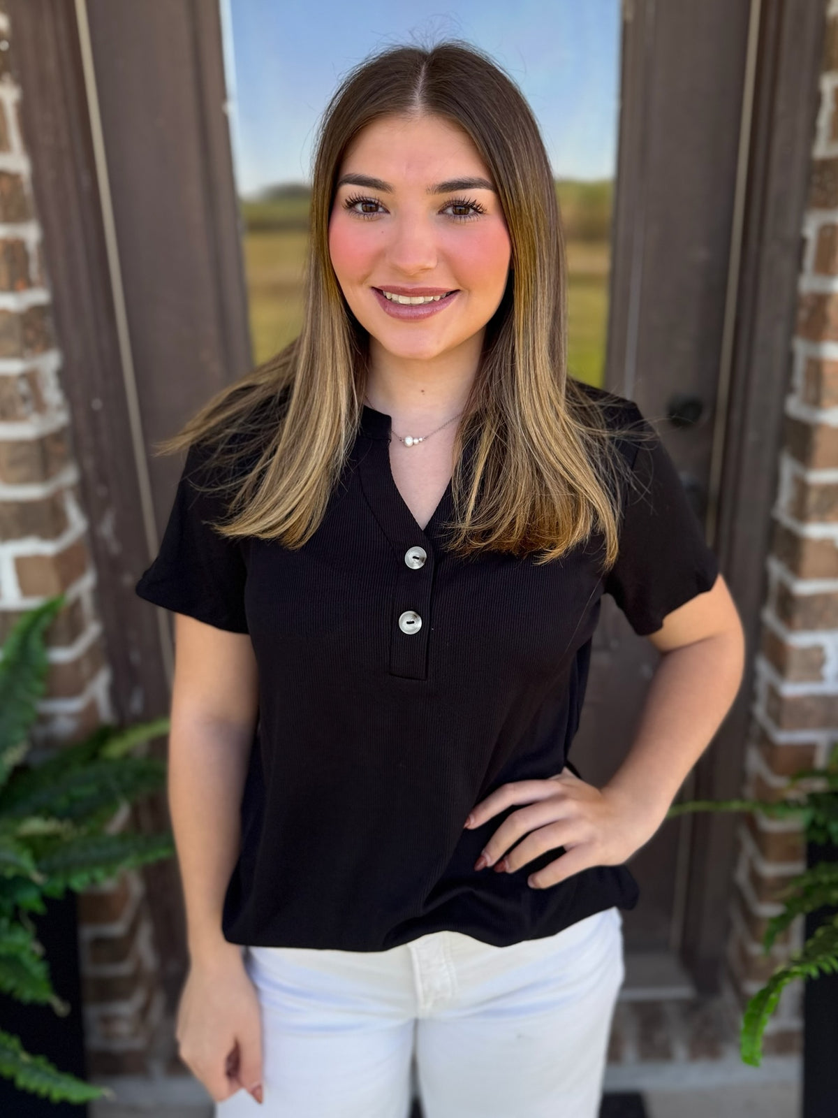 A woman wearing a black short-sleeve henley shirt with buttons and white pants, standing in front of a brick doorway.