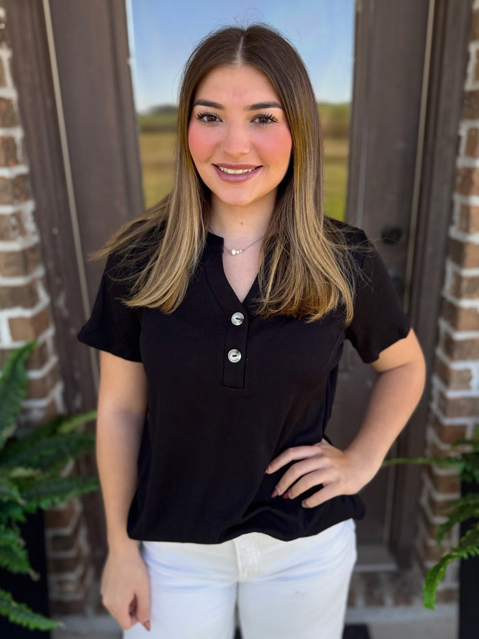 A woman wearing a black short-sleeve henley shirt with buttons and white pants, standing in front of a brick doorway.