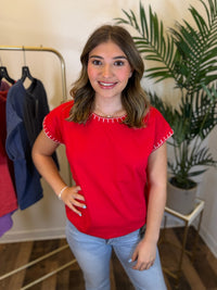 A woman modeling a bright red short-sleeve top with white stitch trim along the neckline and sleeve hems, paired with light blue jeans in an indoor boutique setting.