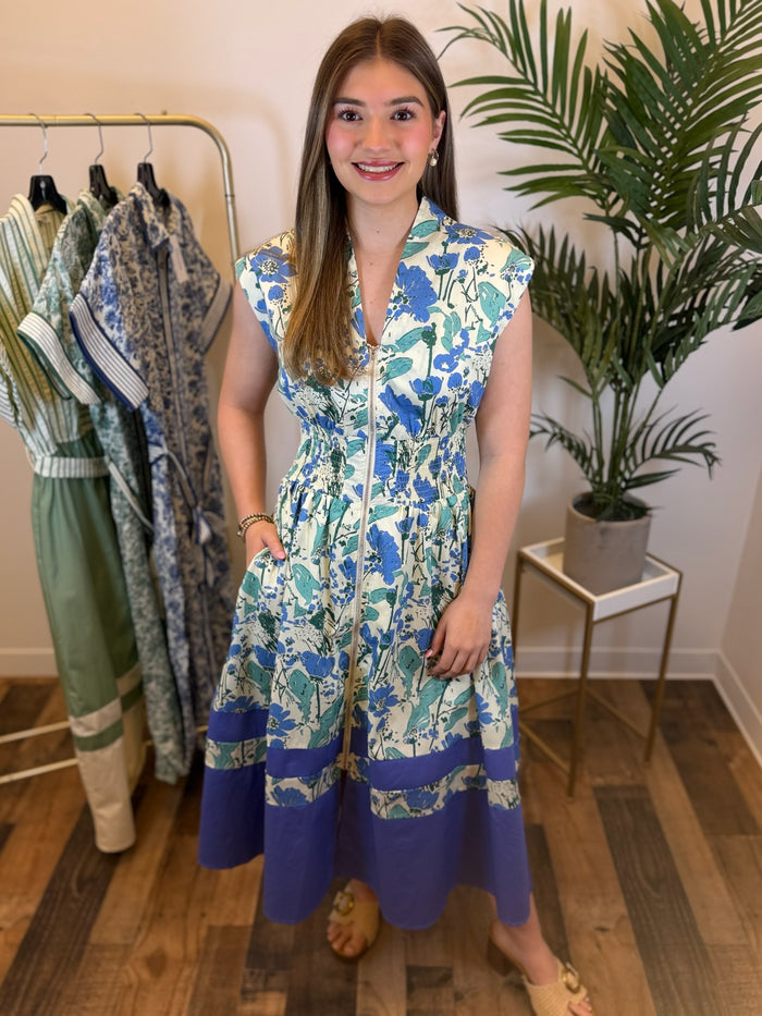 Woman wearing a sleeveless blue and green floral midi dress with zip front and smocked waist standing in a room with clothes and plants.