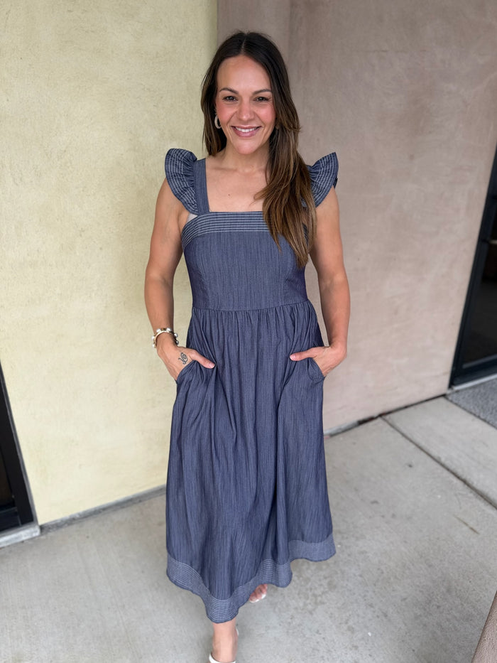 A woman wearing a sleeveless midi dress with ruffled shoulder straps, a square neckline, and side pockets, standing outdoors against a beige stucco wall.
