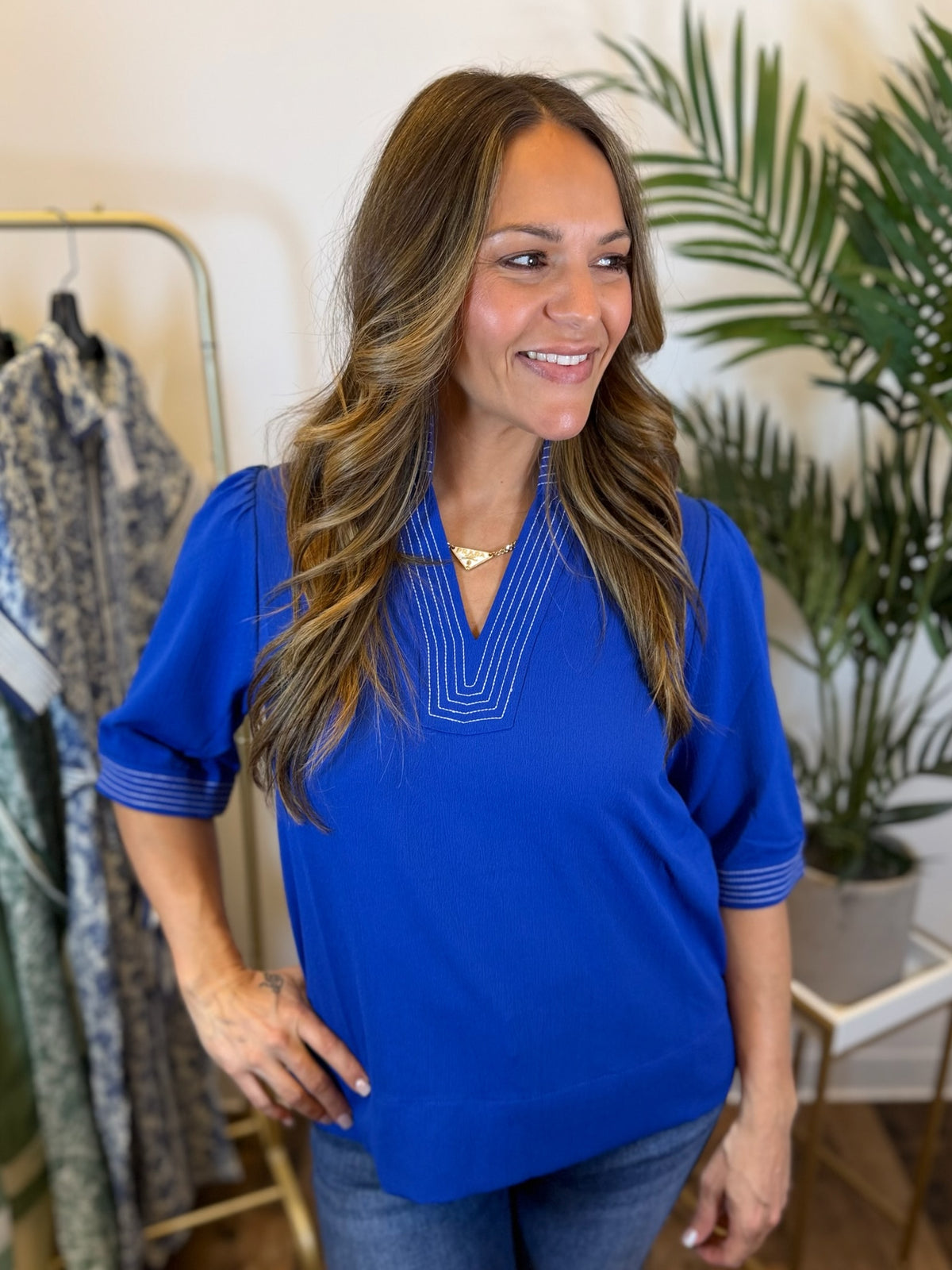 Woman wearing a royal blue top with stitched trim details and 3/4 sleeves in a clothing store with plants in the background