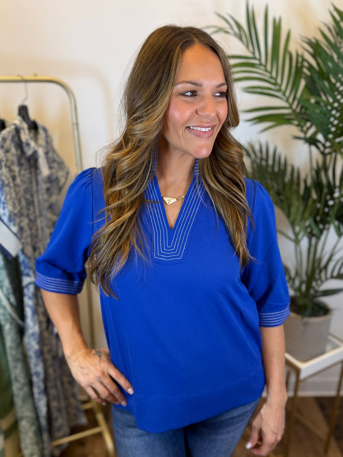 Woman wearing a royal blue top with stitched trim details and 3/4 sleeves in a clothing store with plants in the background