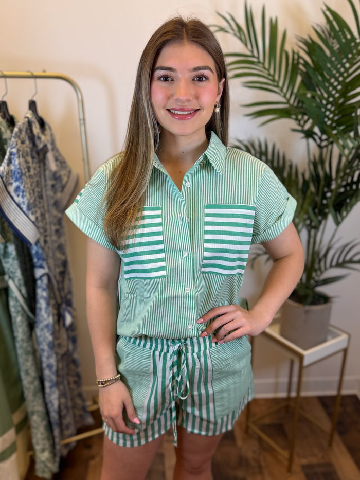 Woman wearing a green and white striped button front top and coordinating shorts with drawstring detail in a room with plants and clothing racks.