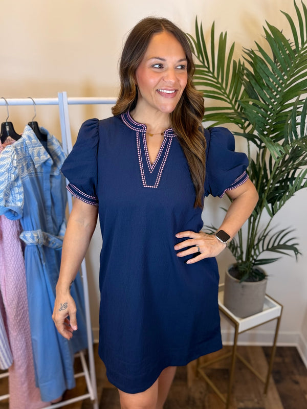 Woman wearing navy puff sleeve dress with split neckline and pink trim details standing in a room with plant and clothing rack.