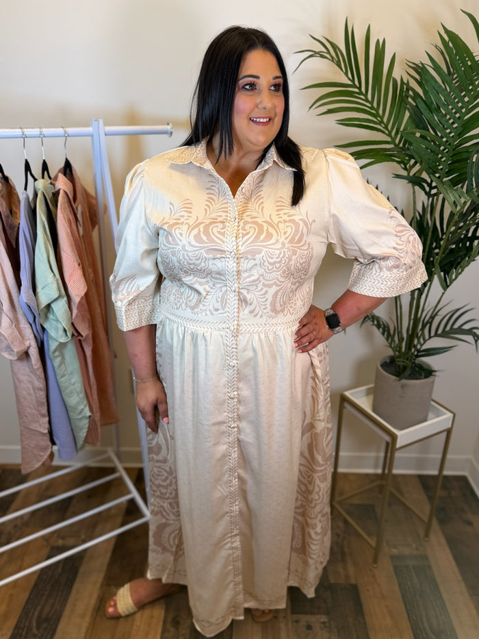 Woman wearing a white dress with intricate patterns in a room with clothing racks and a plant.