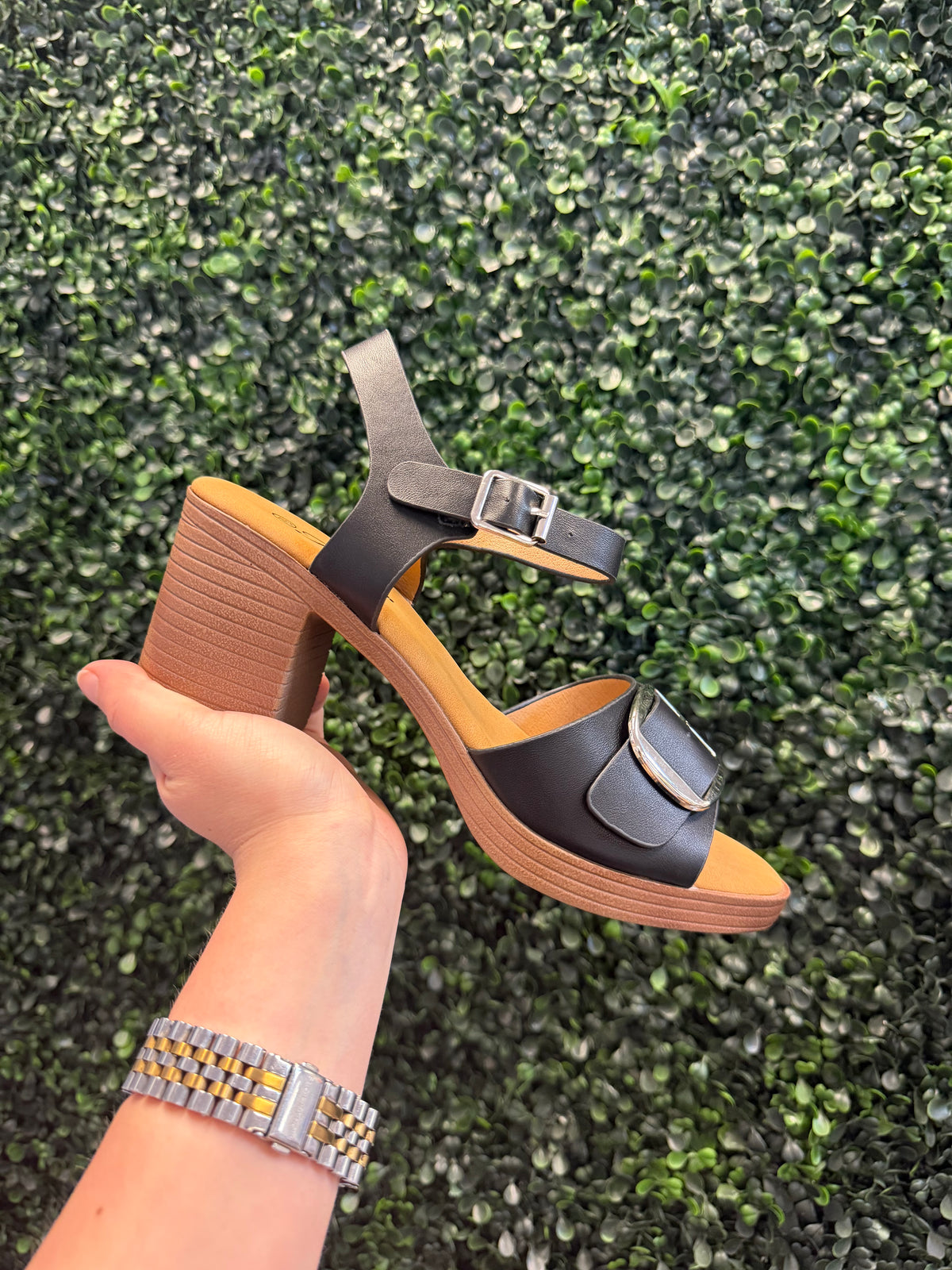 Wooden-heeled shoe with black strap held by a hand against a green textured background
