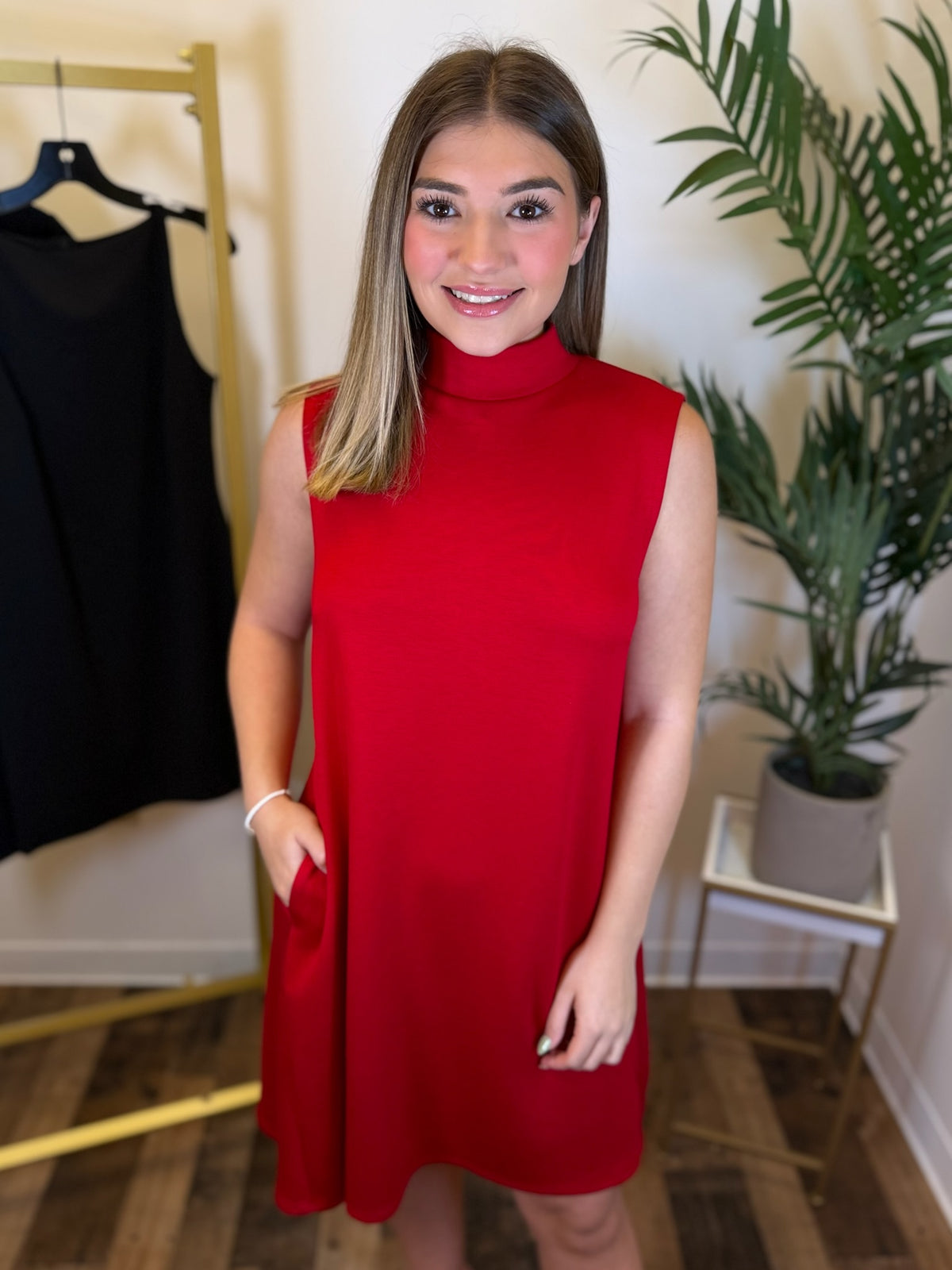 A woman wearing a sleeveless, high-neck red dress with side pockets, standing in a boutique setting with a plant and a clothing rack in the background.