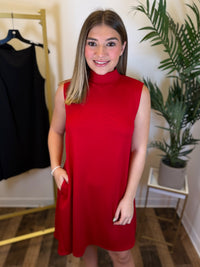A woman wearing a sleeveless, high-neck red dress with side pockets, standing in a boutique setting with a plant and a clothing rack in the background.