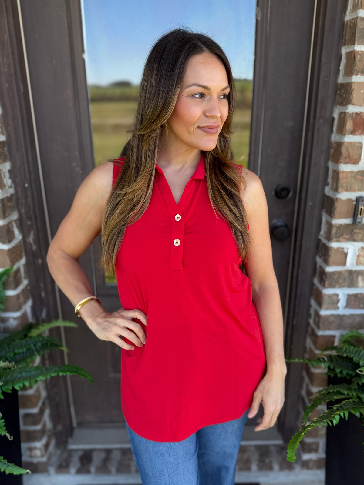 A woman wearing a bright red sleeveless polo shirt with a button placket and blue jeans, standing in front of a brick door with plants visible on either side.
