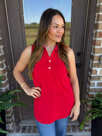 A woman wearing a bright red sleeveless polo shirt with a button placket and blue jeans, standing in front of a brick door with plants visible on either side.