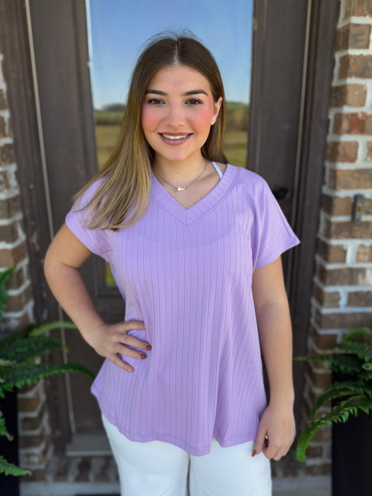 A woman wearing a ribbed lavender V-neck top with short sleeves stands in front of a brick wall.