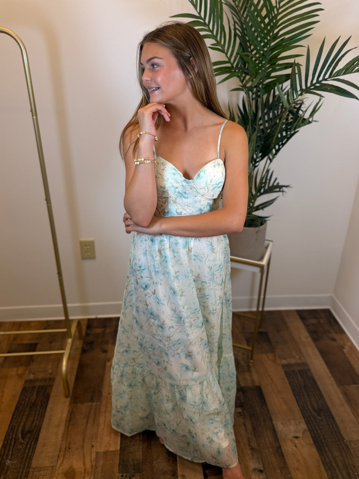 Woman in a floral dress standing in a room with wooden flooring and a plant.