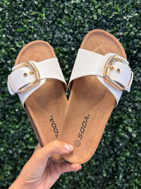 Pair of white slip on sandals with gold buckles held against a green grass background