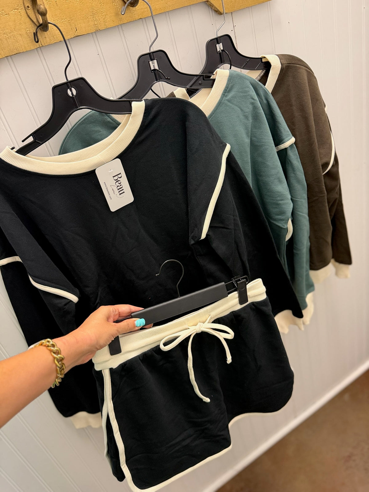 A person's hand holds a black and cream two-piece lounge set hanging on a black hanger against a white paneled wall, with other lounge sets in teal and brown hanging behind it.