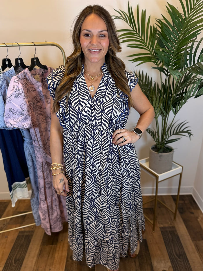 Woman in a cream and navy patterned maxi dress with ruffle sleeves and split neckline standing in a clothing store with plants and racks of clothes in the background.