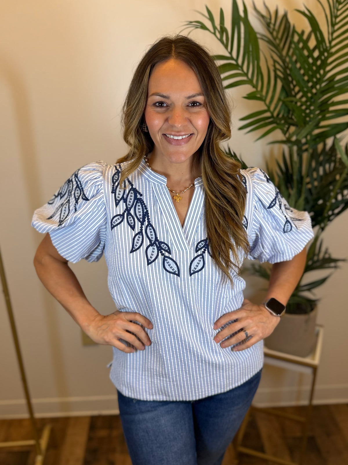 Woman wearing a white and blue striped blouse with puff sleeves and navy embroidered leaf patterns, standing indoors.