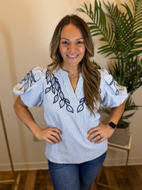 Woman wearing a white and blue striped blouse with puff sleeves and navy embroidered leaf patterns, standing indoors.