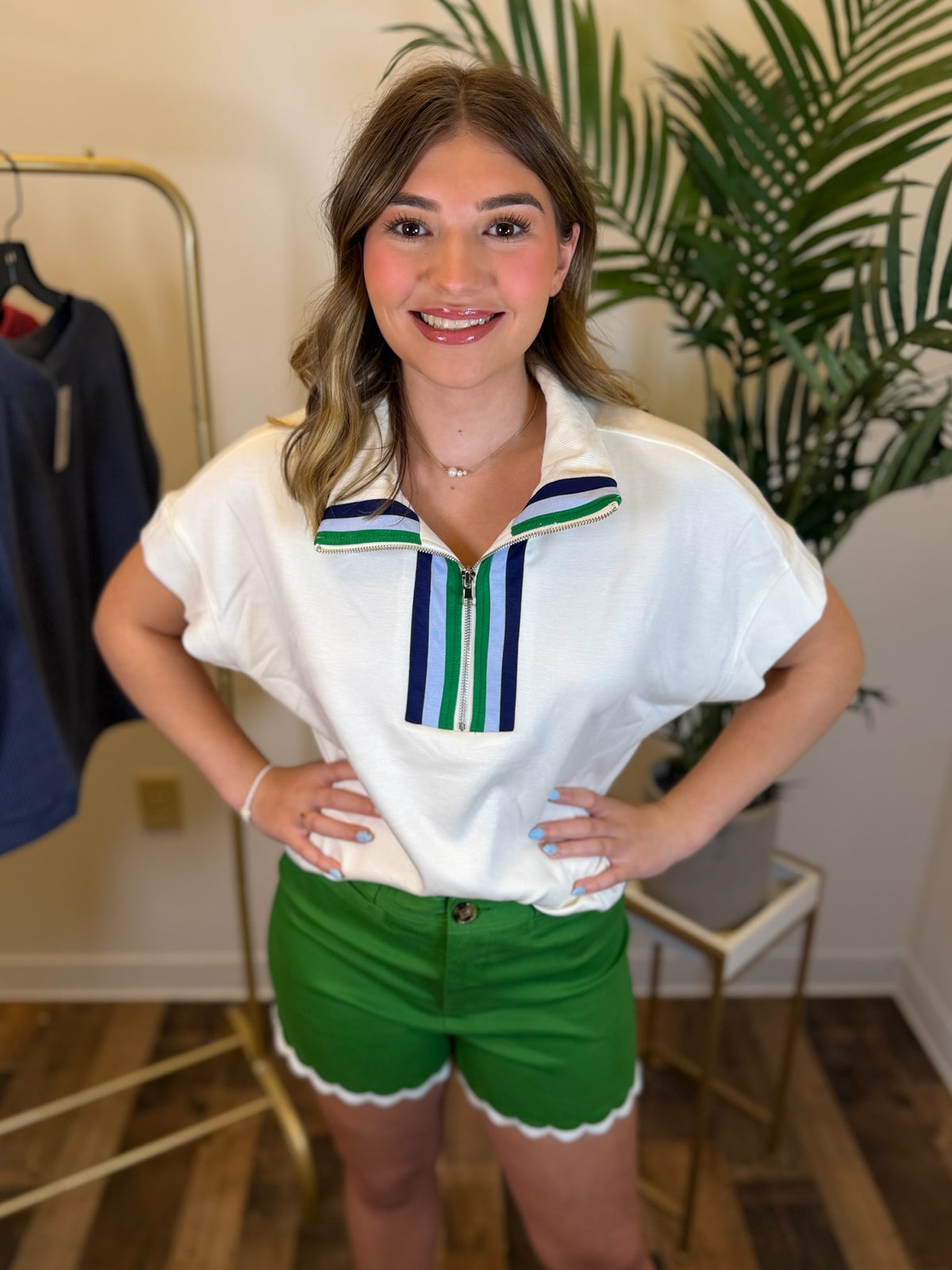 A woman modeling a white short-sleeve polo shirt with green and blue striped collar details, paired with green shorts, standing in an indoor boutique setting.