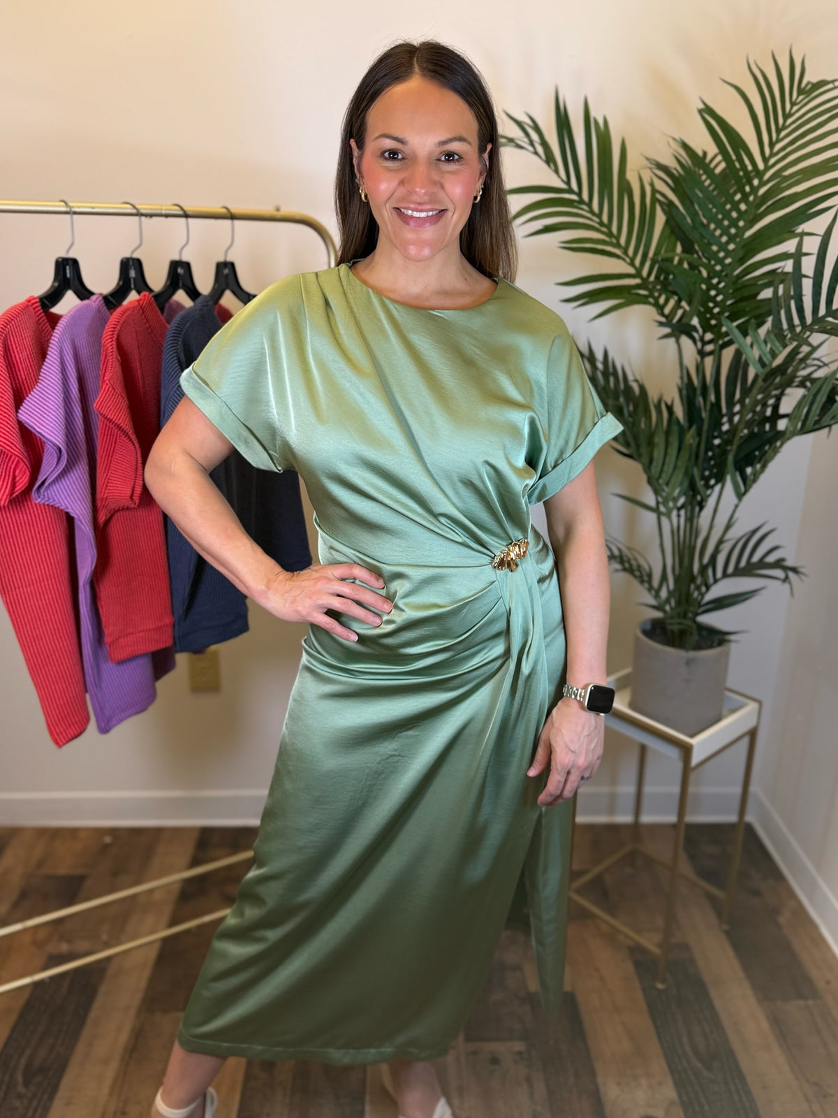 A woman modeling a sage green satin midi dress with short sleeves and a twisted waist detail, standing in a boutique setting with a clothing rack and a potted plant in the background.