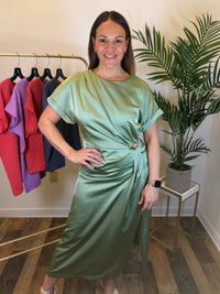 A woman modeling a sage green satin midi dress with short sleeves and a twisted waist detail, standing in a boutique setting with a clothing rack and a potted plant in the background.