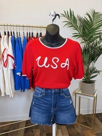 Mannequin wearing a red 'USA' puff sleeve shirt with blue shorts in a store setting.