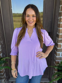 Woman wearing a lavender blouse with short puff sleeves, smocked detailing, and a split V-neckline.
