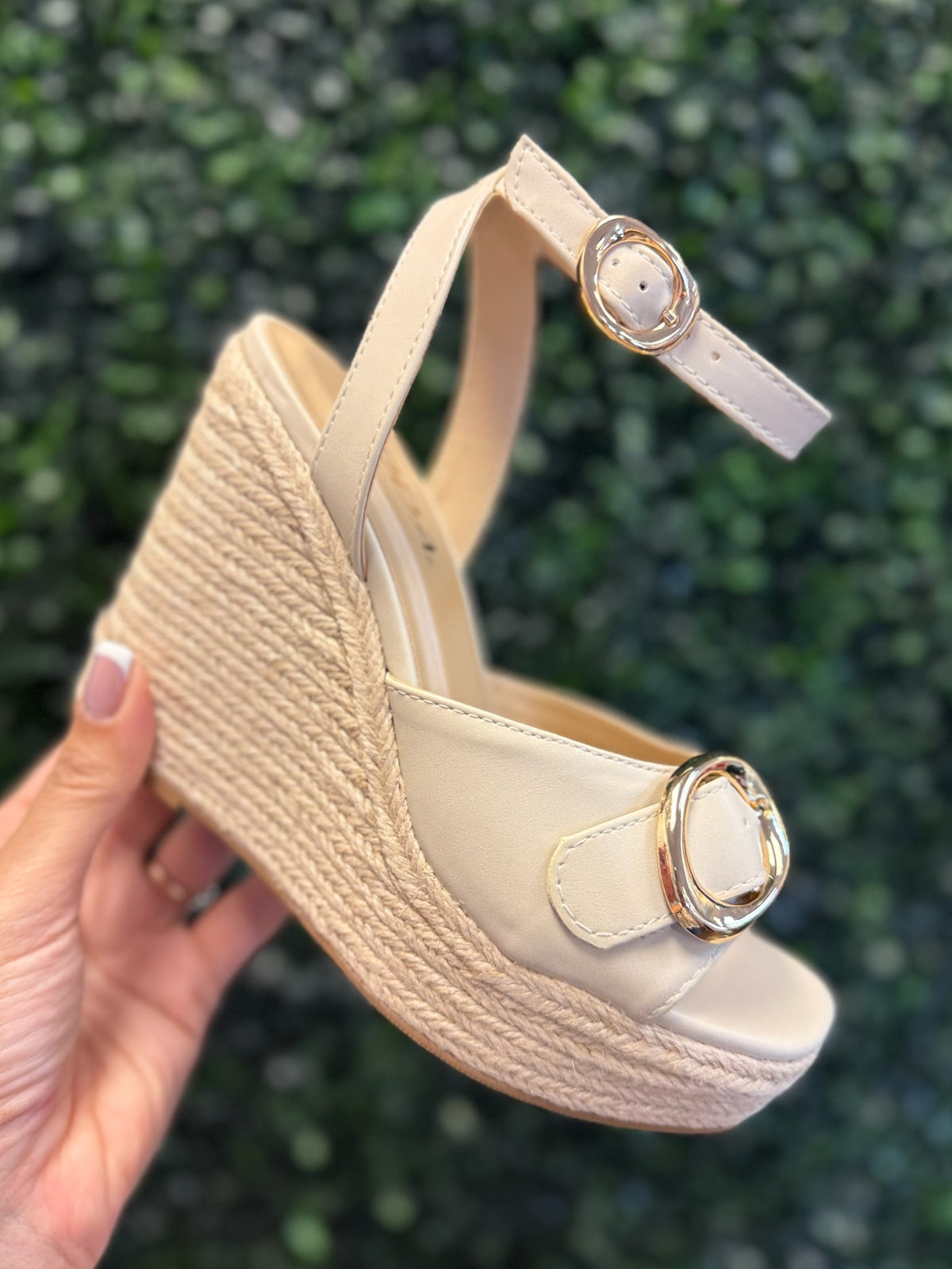 Beige wedge sandal with braided sole held against a green leafy background