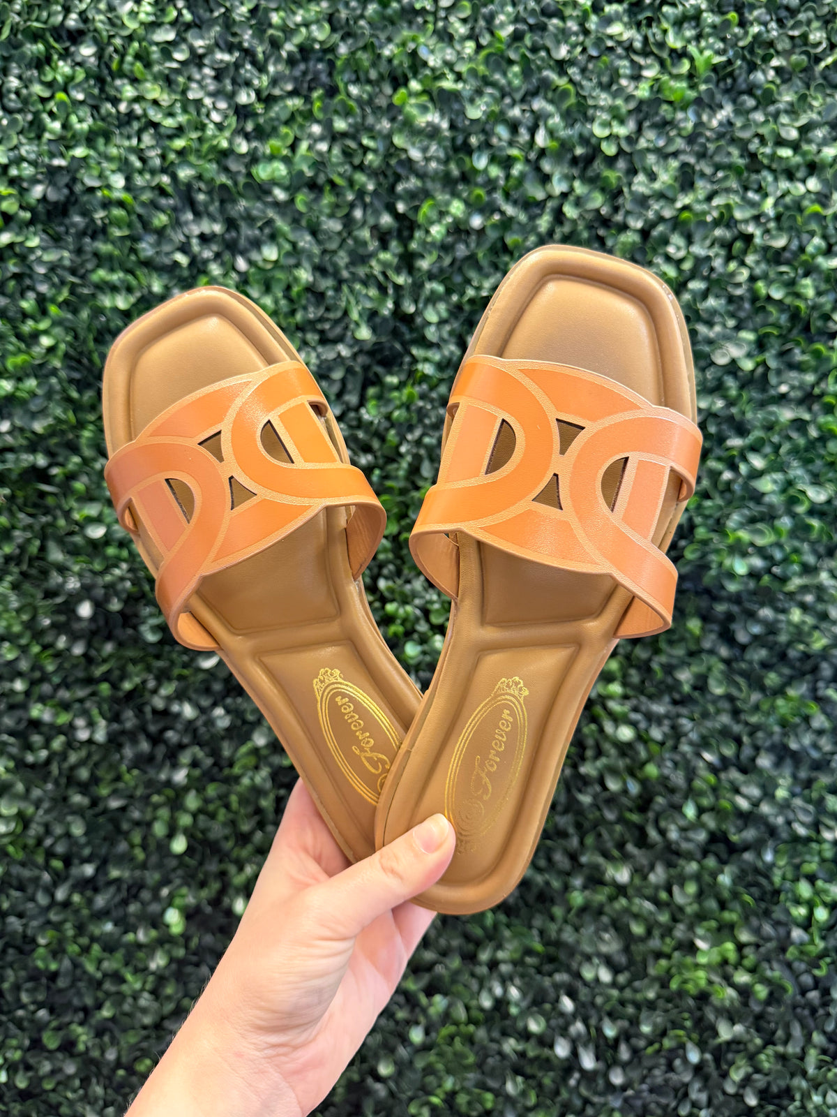 Pair of brown slip on sandals with cutout details held against a green textured background