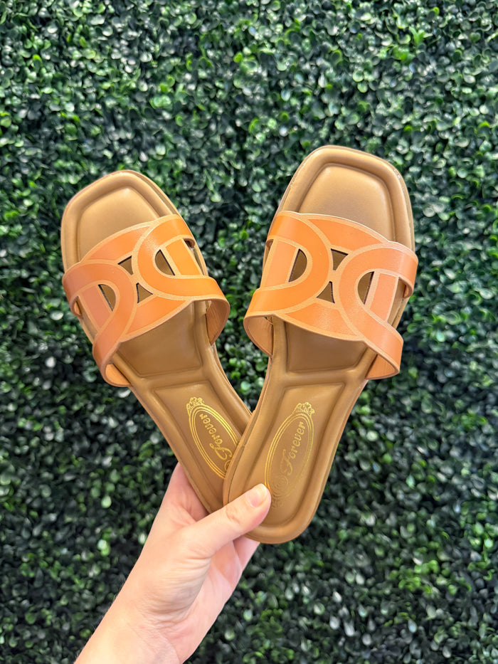 Pair of brown slip on sandals with cutout details held against a green textured background