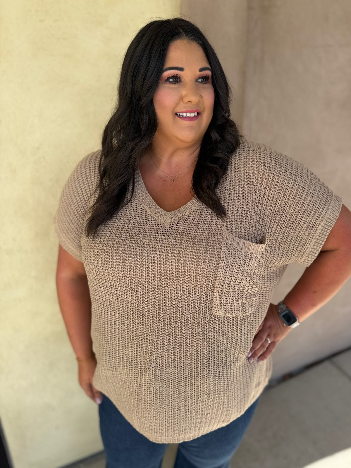 A woman wearing a beige short-sleeve knit V-neck sweater with a chest pocket, paired with blue jeans, standing against a textured beige wall.